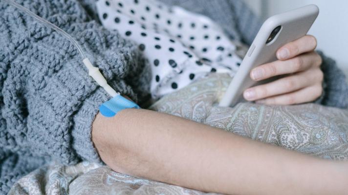 Patient in a blue sweater comforter recuperates at home and receives an IV infusion while checking patient portal information on a cell phone.